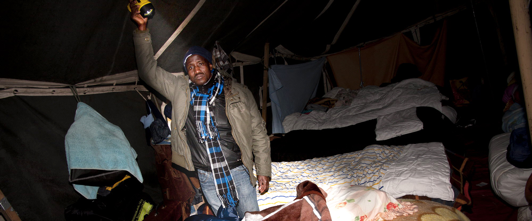 In het schijnsel van zijn zaklamp toont een migrant, die illegaal in Nederland is, zijn slaapplaats in een tentenkamp in Osdorp. | Foto: Erixphotobook | Nationale Beeldbank In het schijnsel van zijn zaklamp toont een migrant, die illegaal in Nederland is, zijn slaapplaats in een tentenkamp in Osdorp. | Foto: Erixphotobook | Nationale Beeldbank